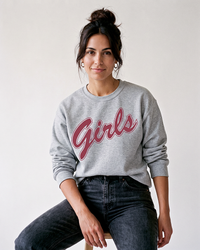 Brunette woman with hair in a messy bun wearing a grey Girls sweatshirt with rose-red script graphic and black jeans.