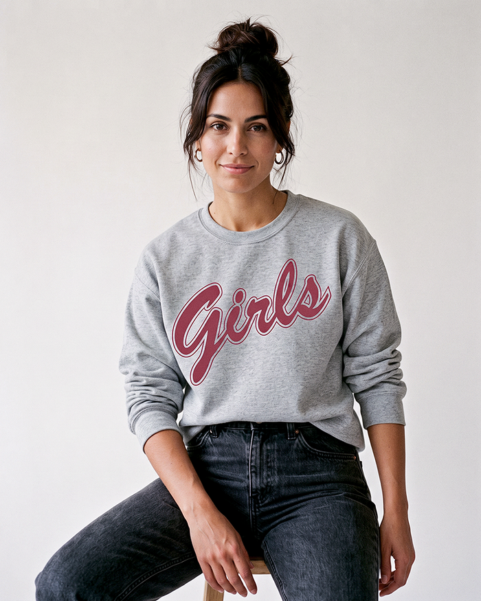 Brunette woman with hair in a messy bun wearing a grey Girls sweatshirt with rose-red script graphic and black jeans.