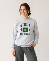 Woman with auburn hair wearing an ash gray Girls Football crewneck sweatshirt with forest green varsity graphic and gray pants.