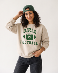 Woman wearing a sand Girls Football crewneck sweatshirt with green football graphic, dark jeans and a backward green baseball cap.