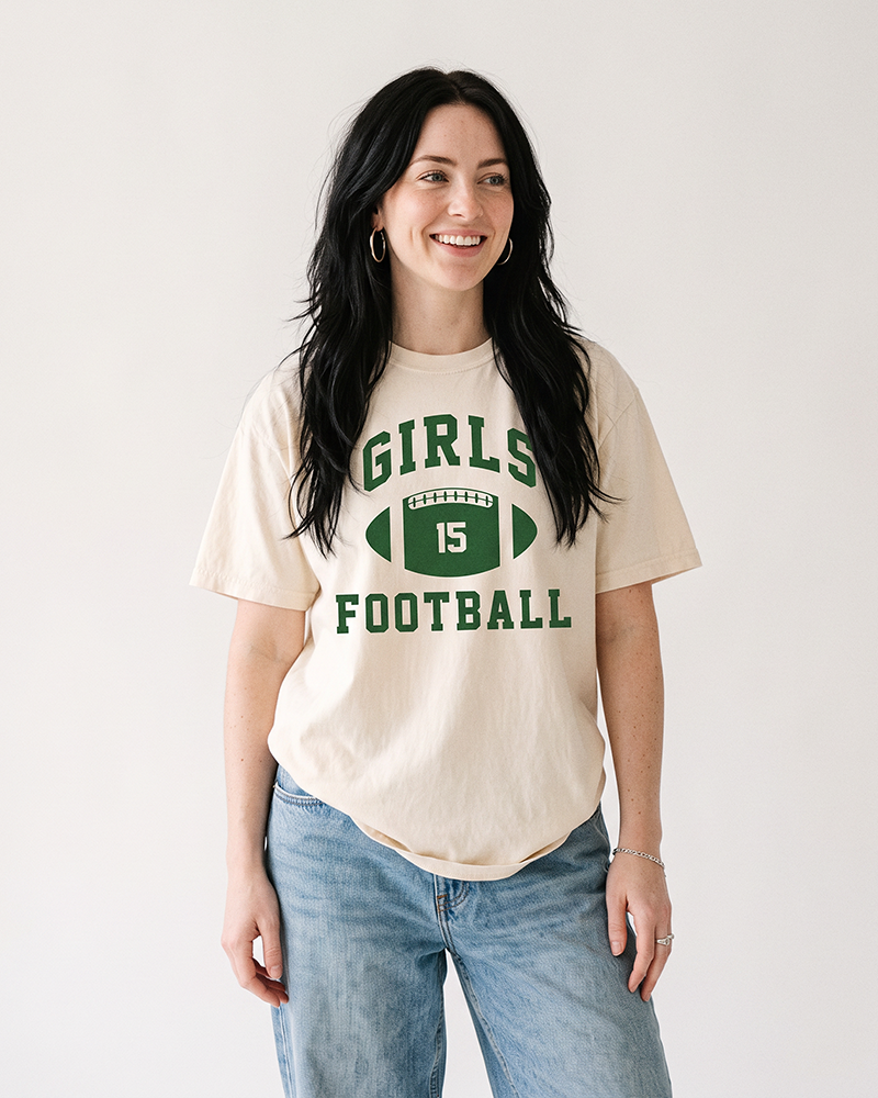 Woman with long dark hair wearing an ivory Girls Football heavyweight graphic tee with green collegiate lettering and blue jeans.