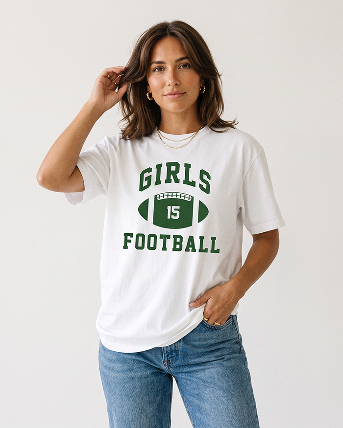 Brunette woman modeling a white Girls Football heavyweight tee with green vintage football graphic and blue jeans.