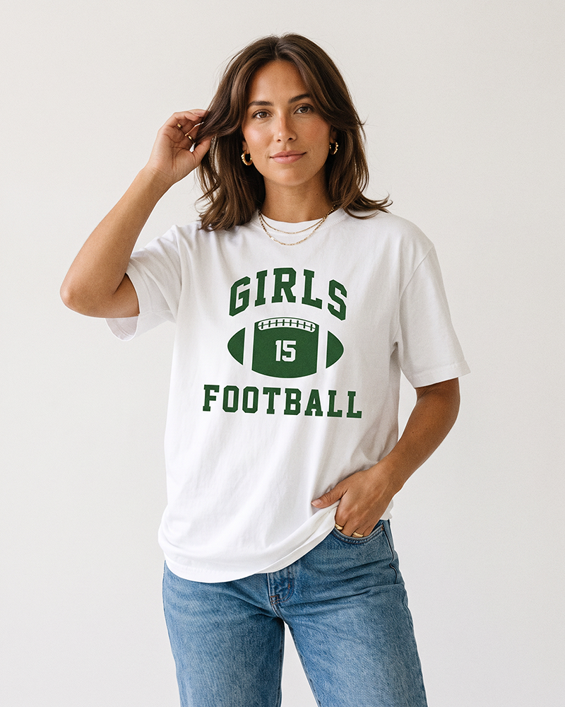 Brunette woman modeling a white Girls Football heavyweight tee with green vintage football graphic and blue jeans.