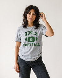 Brunette woman wearing an Athletic Heather Girls Football tee with green collegiate graphic and black jeans.