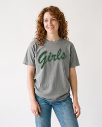 Woman with curly auburn hair wearing a grey Girls heavyweight graphic tee with forest green script lettering and blue jeans.