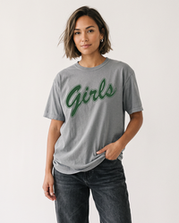 Woman with a short brunette bob wearing a grey Comfort Colors heavyweight Girls graphic t-shirt with green lettering and black jeans.