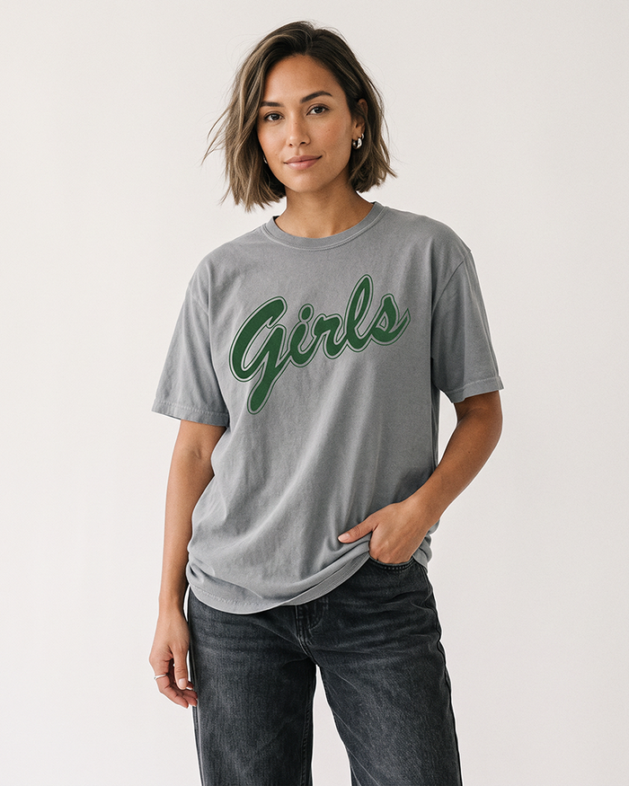 Woman with a short brunette bob wearing a grey Comfort Colors heavyweight Girls graphic t-shirt with green lettering and black jeans.