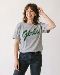 Woman with shoulder-length brown hair wearing an athletic heather grey Girls graphic tee with green script lettering and dark jeans.