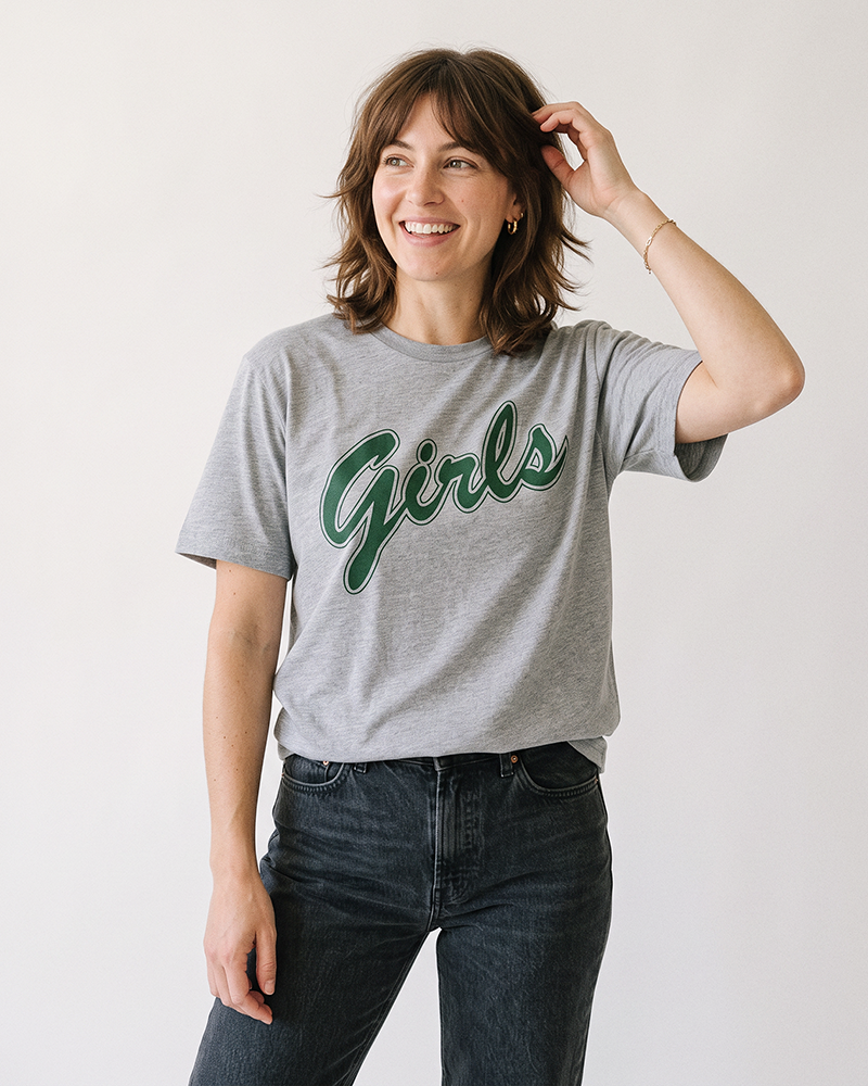 Woman with shoulder-length brown hair wearing an athletic heather grey Girls graphic tee with green script lettering and dark jeans.