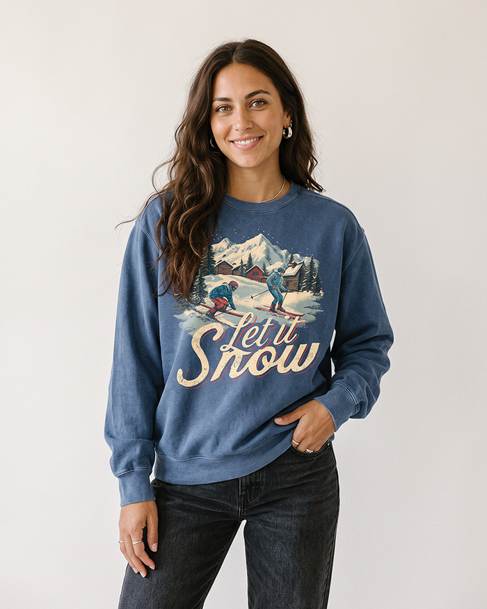 Woman wearing a denim blue Let It Snow heavyweight sweatshirt with vintage skiing and snowy mountain village graphic, styled with black jeans.
