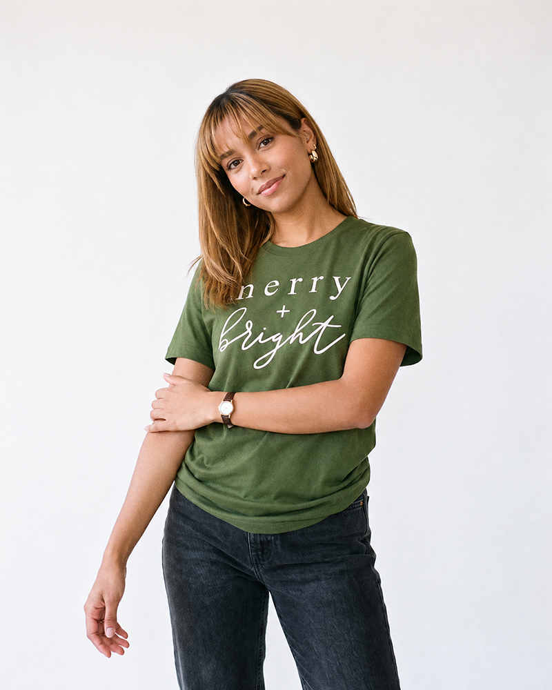 Woman wearing olive Merry + Bright Christmas graphic tee with white lettering