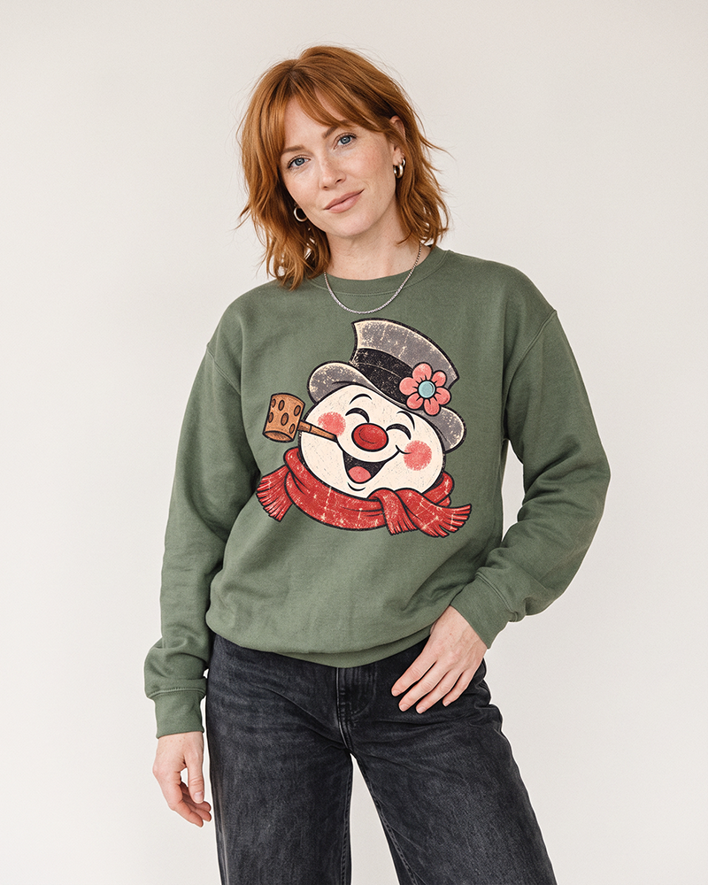 Red-haired woman wearing a Military Green vintage snowman Christmas sweatshirt with dark jeans.