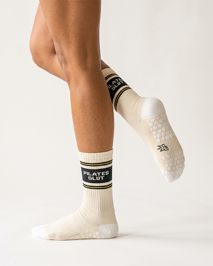 Female model wearing Pilates Slut grip socks with white honeycomb grip pattern visible on raised foot