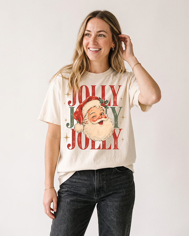 Blonde woman wearing an ivory retro Santa Christmas graphic tee with red and green Jolly Jolly Jolly print and dark denim jeans.