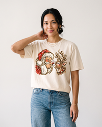 Woman wearing an ivory vintage Santa Christmas graphic tee with a smiling reindeer, festive holiday artwork, and blue jeans.