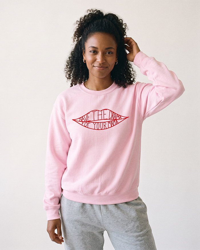 Woman with curly hair wearing a light pink Save The Drama For Your Mama graphic sweatshirt and gray lounge pants.