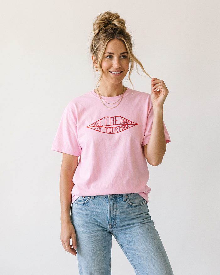 Blonde woman with messy bun modeling a blossom pink Save The Drama For Your Mama graphic tee and light wash jeans.