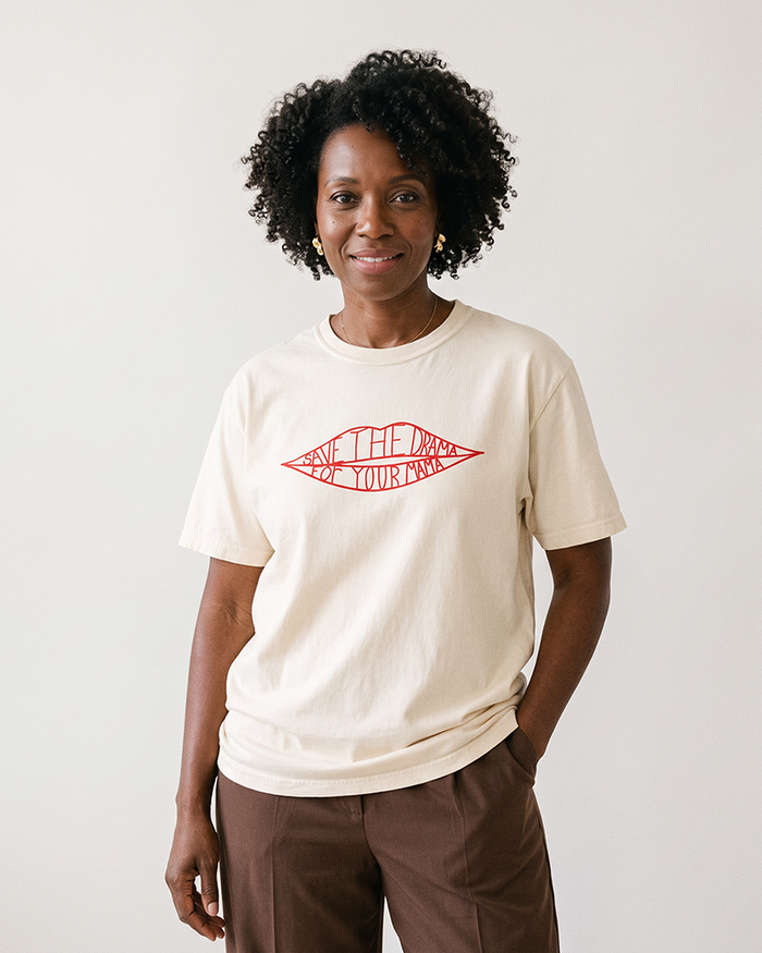 Woman with natural curly hair wearing an ivory Save The Drama For Your Mama graphic T-shirt and brown pants.