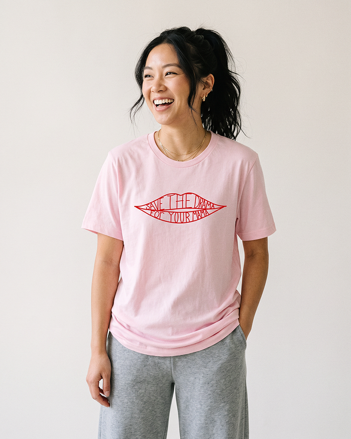 Woman wearing soft pink Save The Drama For Your Mama graphic tee with gray casual pants.