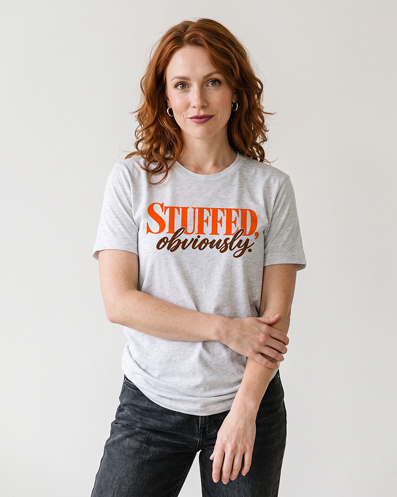 Woman wearing an ash grey Stuffed, Obviously Thanksgiving graphic tee with orange and brown lettering and black denim jeans.