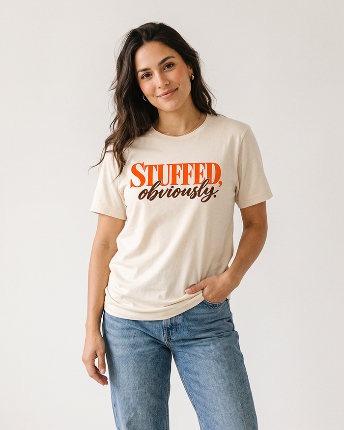 Woman wearing a natural cream Stuffed, Obviously Thanksgiving graphic tee with orange and brown lettering and blue jeans.