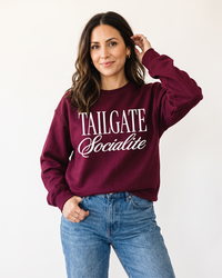 Woman wearing maroon Tailgate Socialite crewneck sweatshirt with white graphic and jeans